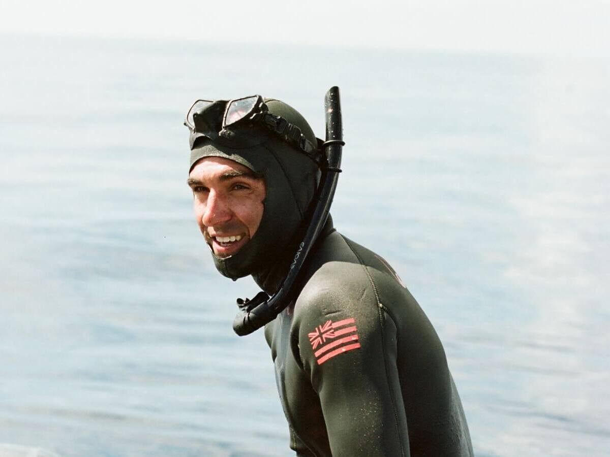 Dylan Currier smiling on the stern of his boat in a hooded wetsuit, mask pushed up on his head, the open ocean behind him