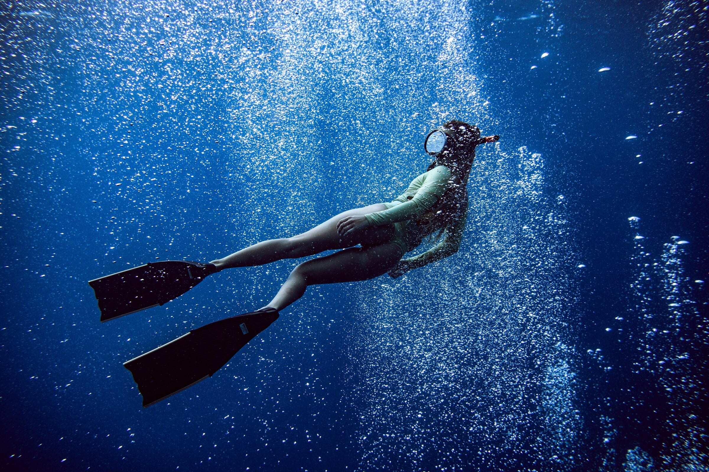 A freediver on long fins rising through a curtain of bubbles in deep blue water