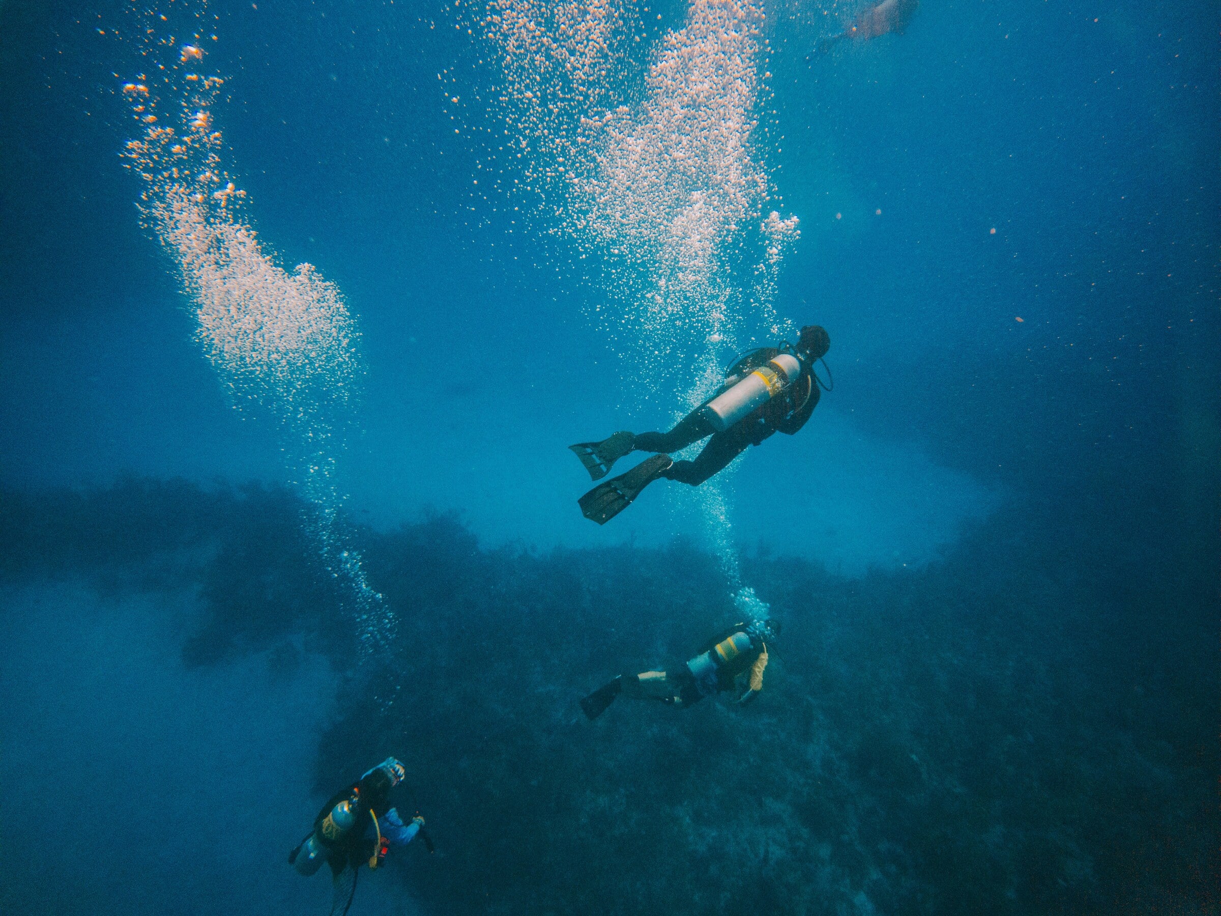 Three scuba divers over a dark reef, columns of bubbles rising above them