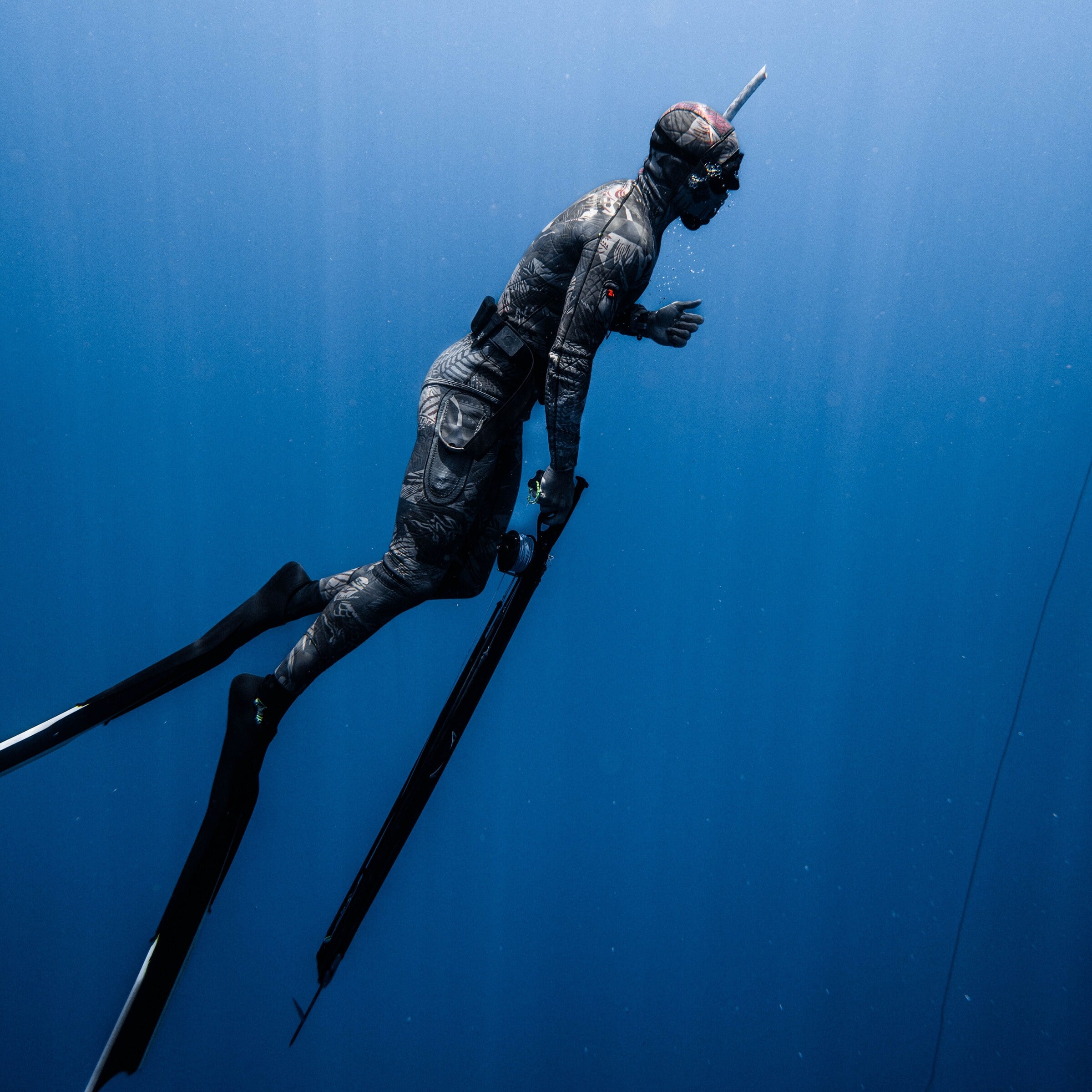 A spearfisher descending through sun rays in deep blue water, speargun along the body